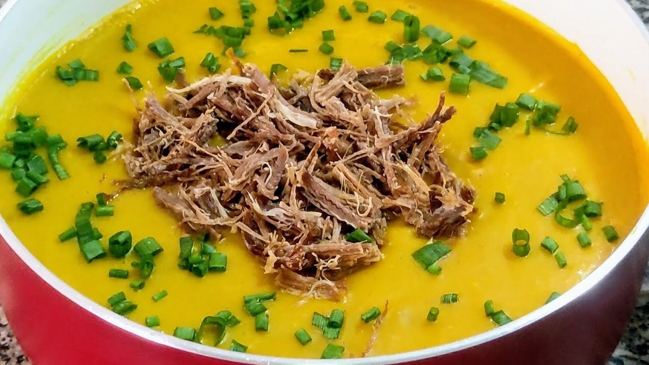 Caldo de abóbora cremoso com carne seca desfiada e cebolinha por cima, servido na panela vermelha