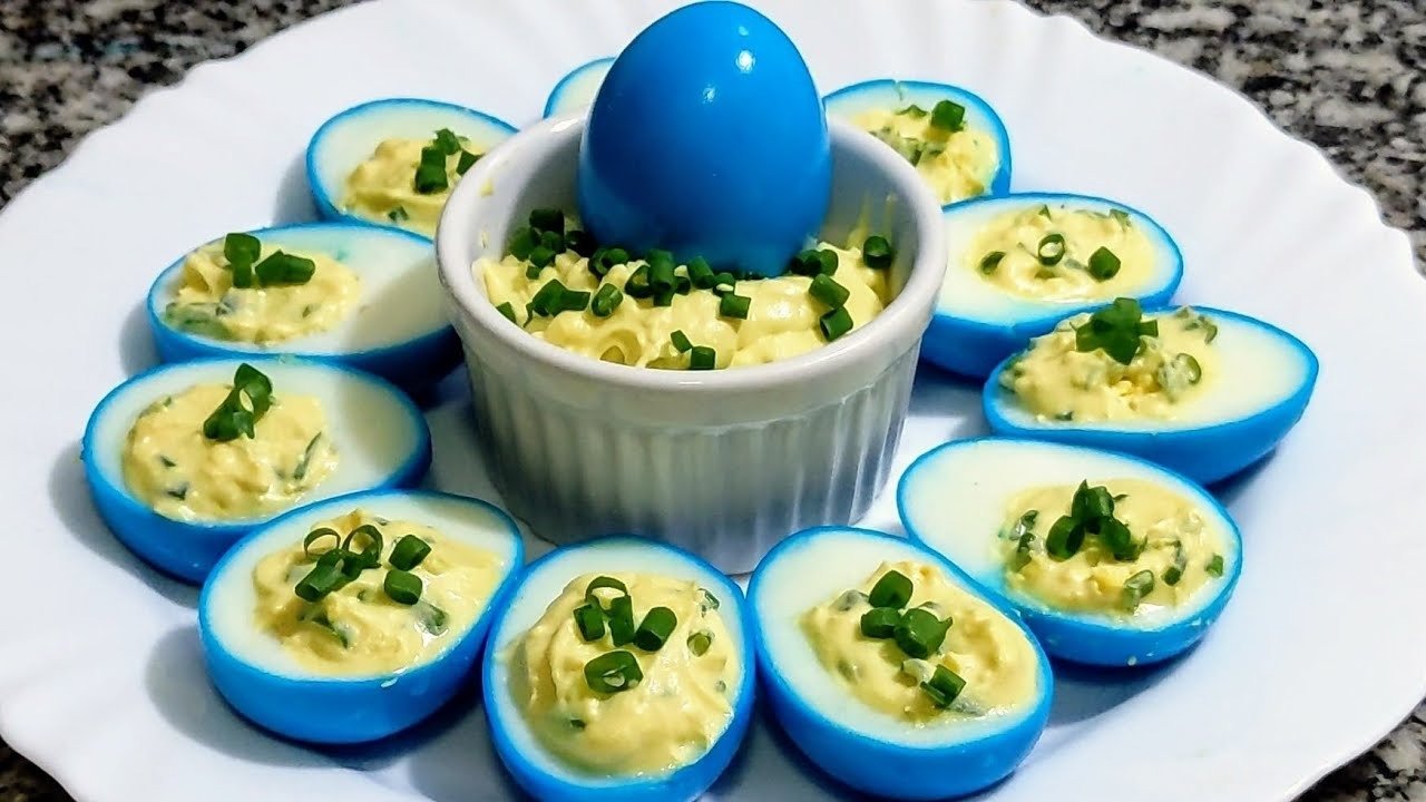 Ovos cozidos tingidos de azul, cortados ao meio e recheados com creme de gema e cebolinha, em prato branco ao redor de um potinho com recheio e um ovo azul inteiro