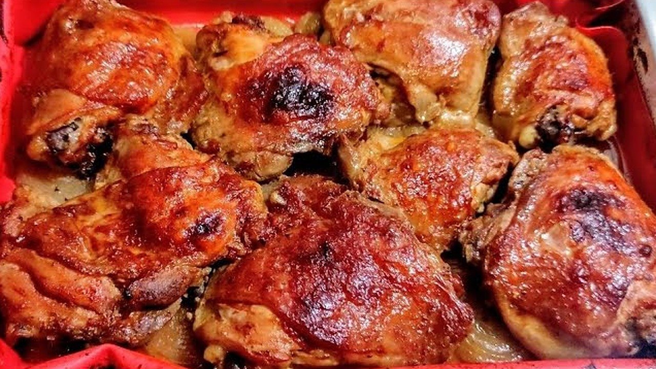Sobrecoxas de frango assadas, douradas e suculentas, em assadeira forrada com tapete culinário vermelho sobre rodelas de cebola